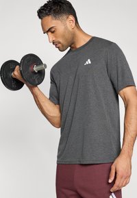 Γκρι αθλητική κοντομάνικη μπλούζα με υφή και ένα μικρό λευκό λογότυπο, σε συνδυασμό με μπορντό παντελόνι γυμναστικής· χέρι κρατάει έναν μαύρο αλτήρα.