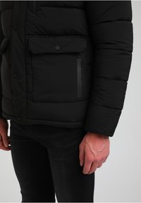 Czarna pikowana kurtka z zapięciem na guzik w kieszeni klapowej oraz kieszenią na zamek, charakteryzująca się pikowanym teksturowaniem i dopasowanym krojem. Nosi się ją na ciemnych dżinsach.