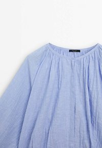 Blouse bleu clair en tissu doux et texturé. Présente un col rond, des manches amples et un détail froncé sur le devant.