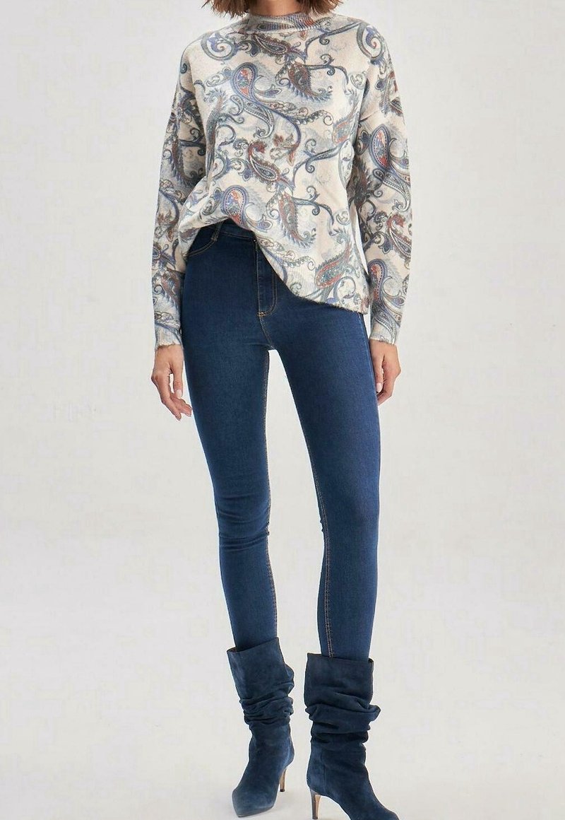 Beige Pullover mit blauem und rotem Paisleymuster; kombiniert mit figurbetonten dunkelblauen Jeans und kniehohen, lässigen blauen Stiefeln.
