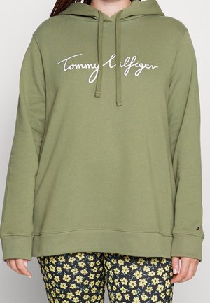 Personne portant un sweat à capuche olive Tommy Hilfiger avec un texte de logo blanc et un pantalon noir à motif floral jaune, visible du cou aux hanches.