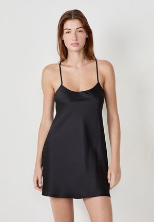 Femme aux longs cheveux bruns portant une robe nuisette en satin noir à fines bretelles, se tenant devant un fond clair simple.