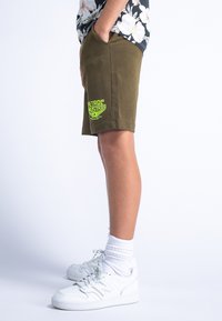 Shorts vert olive à coupe décontractée, arborant un logo imprimé vert néon sur la jambe gauche. Associés à des baskets blanches et des chaussettes texturées.