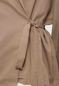 Top maro din țesătură legat în talie cu un nod în formă de fundă, arătând material texturat și mâneci lungi.