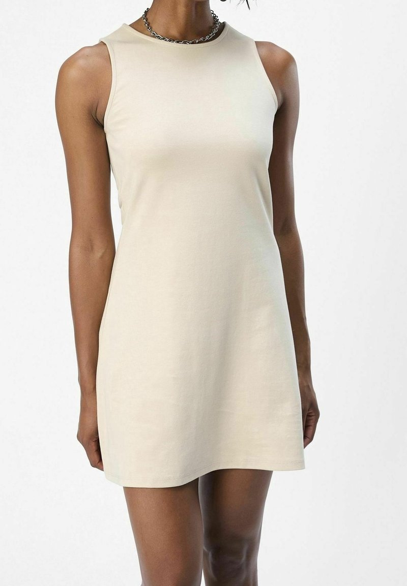 Femme vêtue d'une mini-robe beige sans manches et d'un collier chaîne argenté, debout devant un fond blanc uni.
