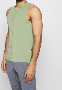 Ljusgrön ärmlös linne gjord av mjukt tyg, med en centrerad söm och subtila sidoklipp, parat med gråa träningsbyxor.