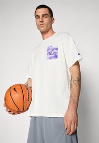 White cotton T-shirt with a purple and white graphic pocket. Loose fit, short sleeves. Holding an orange basketball with black accents.