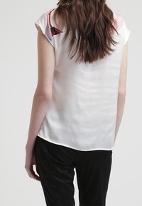 Krátký rukáv, bílá saténová blůza s kulatým výstřihem. Má hladkou texturu a mírně zaoblený lem. Minimalistický design.