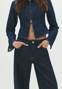 Person wearing dark blue denim jacket zipped halfway and matching high-waisted jeans with hands in pockets, showing a small midriff.