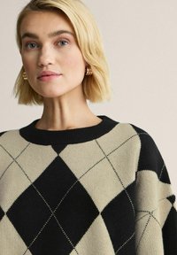 Stickad tröja med ett beige och svart argyle-mönster, rund halsringning och mjuk textur. Gyllene hoops örhängen synliga.