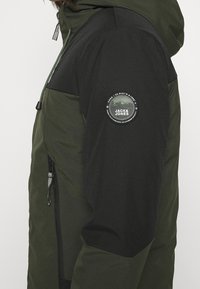 Grön och svart jacka med huva, sidoventil och en rund logotypapplikation på överarmen. Materialet ser slätt ut med en matt finish.