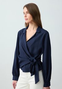 Blusa avvolgente blu navy in tessuto leggero, con maniche lunghe, vita annodata e scollatura a V profonda. Texture liscia, dettagli minimi.
