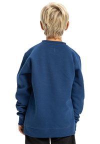 Modrá mikina vyrobená z mäkkej látky, ktorá má voľný strih, manžety s rebrovaním a malú nášivku na zadnej strane.