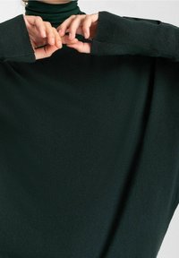 Ciemnozielony sweter dziany o luźnym kroju, z szerokimi ściągaczami i teksturowanym materiałem. Posiada wysoki dekolt, warstwowy pod spód.