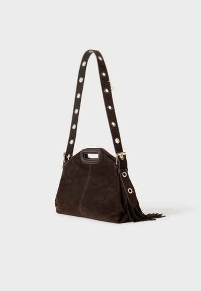 Sac à main en daim marron foncé avec une large bandoulière ornée d'œillets dorés et de détails à pompons, sur un fond uni clair.