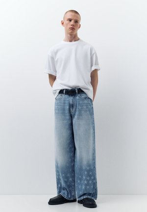 Fiatal férfi fehér, feltűrt ujjú pólóban, széles szárú, kifakult kék farmernadrágban csillagmintákkal, fekete övvel és fekete cipőben, egyszínű fal előtt állva.