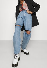 Ljusblå denimjeans med slitna knä utskärningar, hög midja och avslappnad passform, parat med vita snörstövlar och en svart blazer.