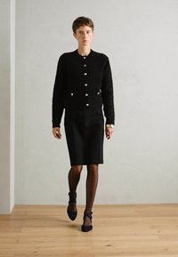 Czarny, puszysty kardigan z zapięciem na guziki, zestawiony z dopasowaną czarną spódnicą. Modelka nosi czarne rajstopy i buty wsuwane. Neutralne tło.