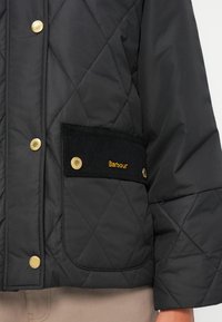 Zwarte gewatteerde jas met gouden hardware, een corduroy pocket met "Barbour" borduurwerk, en een gladde, waterdichte stoftextuur.