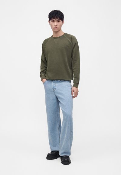 Jeune homme debout portant un pull à manches longues vert olive, un jean bleu clair à jambes larges et des chaussures noires épaisses sur fond blanc.