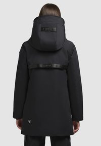 Person in einem schwarzen kapuzenmantel mit weitem Kragen und horizontalem Markenband über dem oberen Rücken, stehend vor einem schlichten Hintergrund.