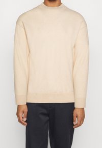 Suéter de punto beige con un cuello redondo alto, mangas largas y puños y dobladillo acanalados. Textura suave; ajuste casual.