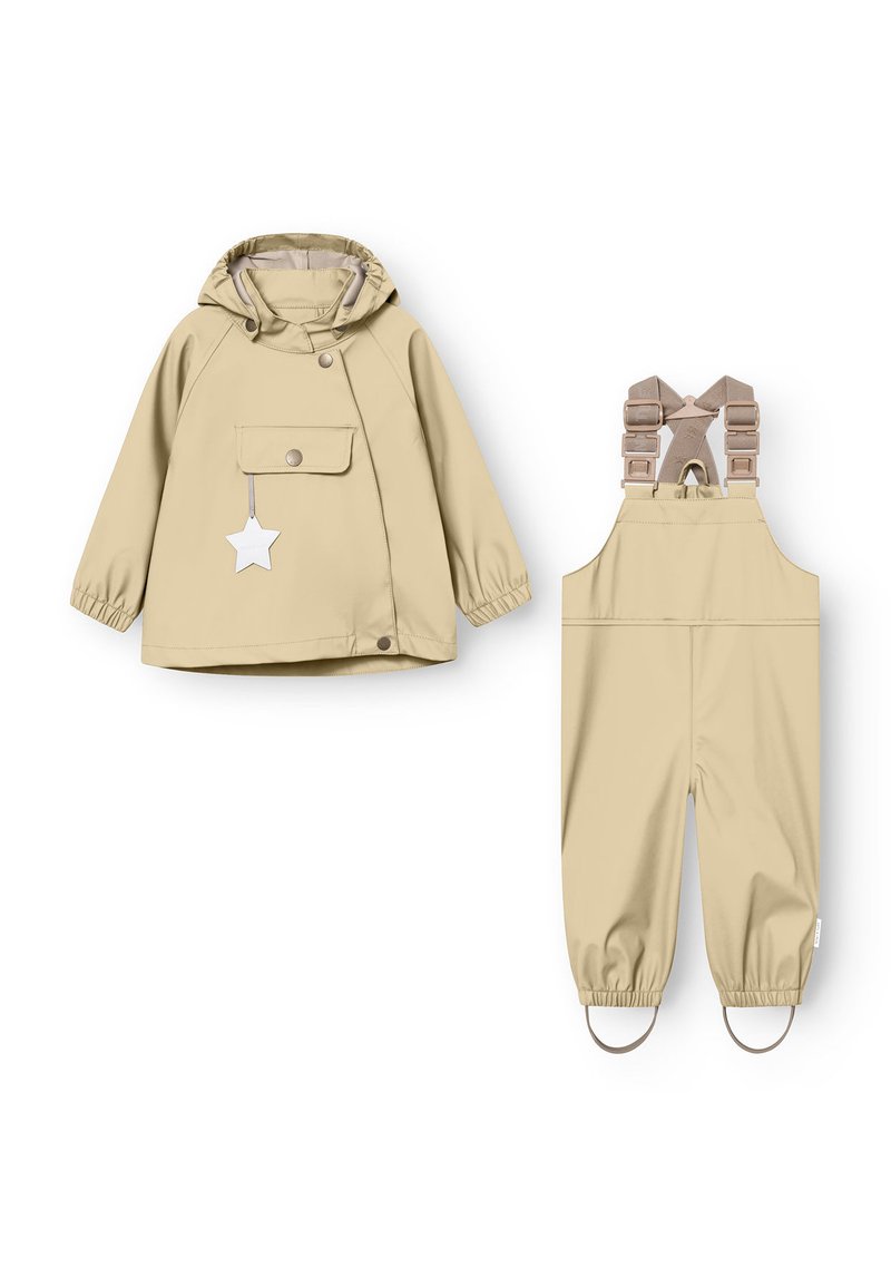 Giacca impermeabile beige con cappuccio, tasca frontale e chiusura a scatto, abbinata a salopette beige con spalline regolabili e polsini elasticizzati.