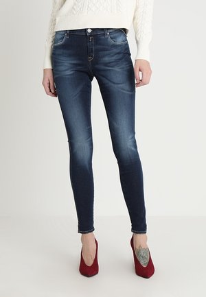 Kvinde iført mørkeblå skinny jeans, hvid striktrøje, røde højhælede sko og en blomster-tatovering på venstre fod.