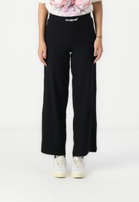 Black wide-leg pants made from soft fabric, featuring an elastic waistband with a white logo. Worn with light-colored sneakers.
