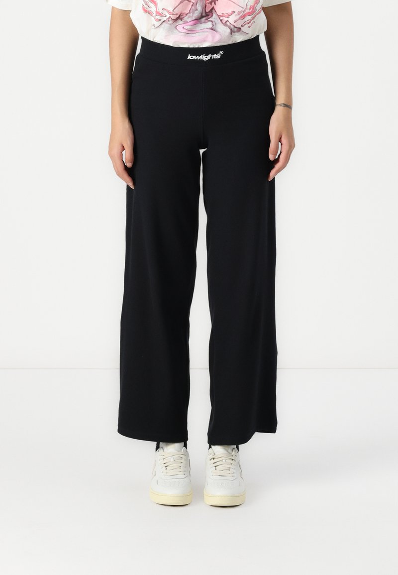 Black wide-leg pants made from soft fabric, featuring an elastic waistband with a white logo. Worn with light-colored sneakers.