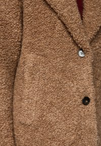 Brauner Textur-Mantel aus einem weichen, flauschigen Stoff, mit einem Reverskragen und zwei Knöpfen – einer schwarz und einer silber. Inklusive einer seitlichen Tasche.