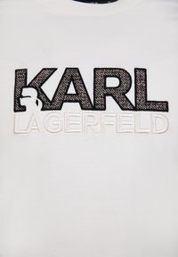 Camiseta blanca de algodón con un texto bordado en negro y gris que dice "KARL LAGERFELD" en negrita, con una silueta de perfil encima del nombre.