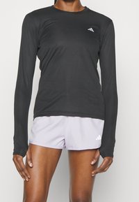 Musta pitkähihainen urheilupaita, jossa on pyöreä kaula-aukko, valmistettu sileästä kankaasta ja jossa on pieni valkoinen logo vasemmassa rinnassa.