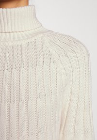Maglione chunky con collo alto, caratterizzato da una trama a costine e motivi a maglia verticale. Primo piano del dettaglio sulla spalla, materiale morbido e leggero.