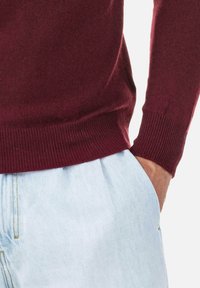 Bordeauxroter Strickpullover mit gerippten Bündchen, kombiniert mit hellblauen, plissierten Denim-Hosen. Fokus auf kontrastierende Texturen und Farben.