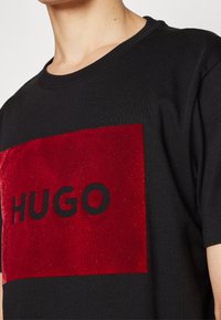 Černé tričko s červeným sametovým obdélníkovým panelem, na kterém je slovo "HUGO" tučně vytištěno černými písmeny. Krátké rukávy, design s kulatým výstřihem.