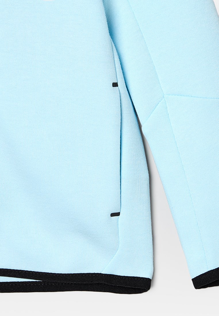 Haut à manches longues bleu clair en tissu texturé, avec des accents noirs à l'encolure et sur le côté. Le devant montre une vue partielle de la manche et de la couture.