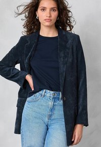 Kvinna med lockigt brunt hår som bär hoopörhängen, en marinblå kordflodblazer, svart skjorta och ljusblå höga jeans mot en grå bakgrund.