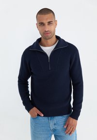 Suéter de punto azul marino con cuello alto y media cremallera. Patrón texturizado en el pecho, puños y dobladillo de canalé. Lleva puesto sobre una camisa blanca.