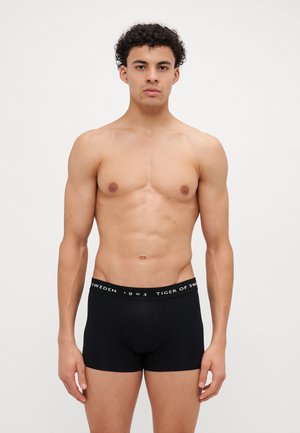 Schwarze Boxershorts mit glatter Textur und elastischem Bund, mit aufgedrucktem Markennamen und Jahr in Weiß. Schlichtes, körperbetontes Design.