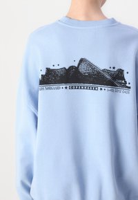 Lys blå hettegenser med rund hals, med et svart grafisk design på baksiden som inkluderer en bygning skisse og tekst detaljer.