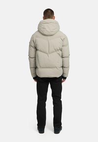 Grå pufferjacka med huva, med en quiltad design och elastiska ärmslut, bärs över svarta byxor och mörka skor, sedd bakifrån.
