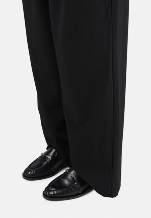 Mocassins en cuir noir avec détail chaîne argentée portés avec un pantalon noir ample sur un fond clair uni.