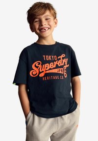 Must mustreest T-särk mustast puuvillast, millel on oranž "Superdry" tekst ja "Tokyo" disain. Kandmine heledate hallide dressipükstega. Loodud vabaks istumiseks, lühikesed varrukad.