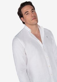 Camisa de lino blanca con cuello abotonado, que presenta botones claros y un sutil bordado en el pecho. Textura suave, estilo casual.