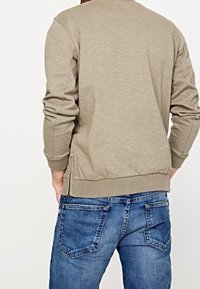 Sudadera a collo alto khaki con polsini e orlo a coste, abbinata a jeans blu che mostrano tasche posteriori e texture in denim.
