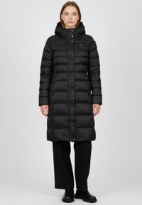 Piumino nero con cappuccio, dal design trapuntato, con bottoni a pressione e lunghezza fino al polpaccio. Indossato sopra pantaloni neri a gamba larga e stivali.