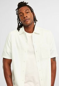Korte mouwen, geknoopte blouse van gestructureerde witte stof met een opgestikte zak. Draag met een effen wit t-shirt. Natuurlijke huidskleur zichtbaar op de arm.
