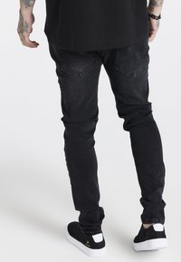 Černé džíny s taperovaným střihem, vybavené dvěma zadními kapsami s diamantovým prošíváním, spárované s černými teniskami s bílými akcenty.