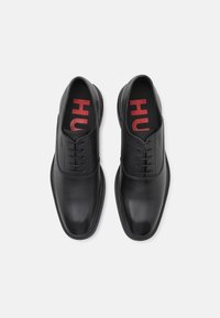 Chaussures habillées en cuir noir avec surfaces lisses et texturées, présentant un design traditionnel à lacets et un logo "HU" rouge sur les semelles intérieures.